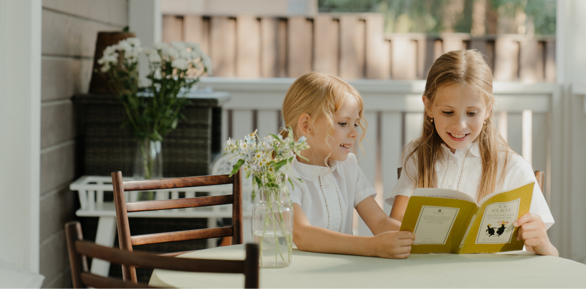 Two young girls reading a book together outdoors on a sunny day.